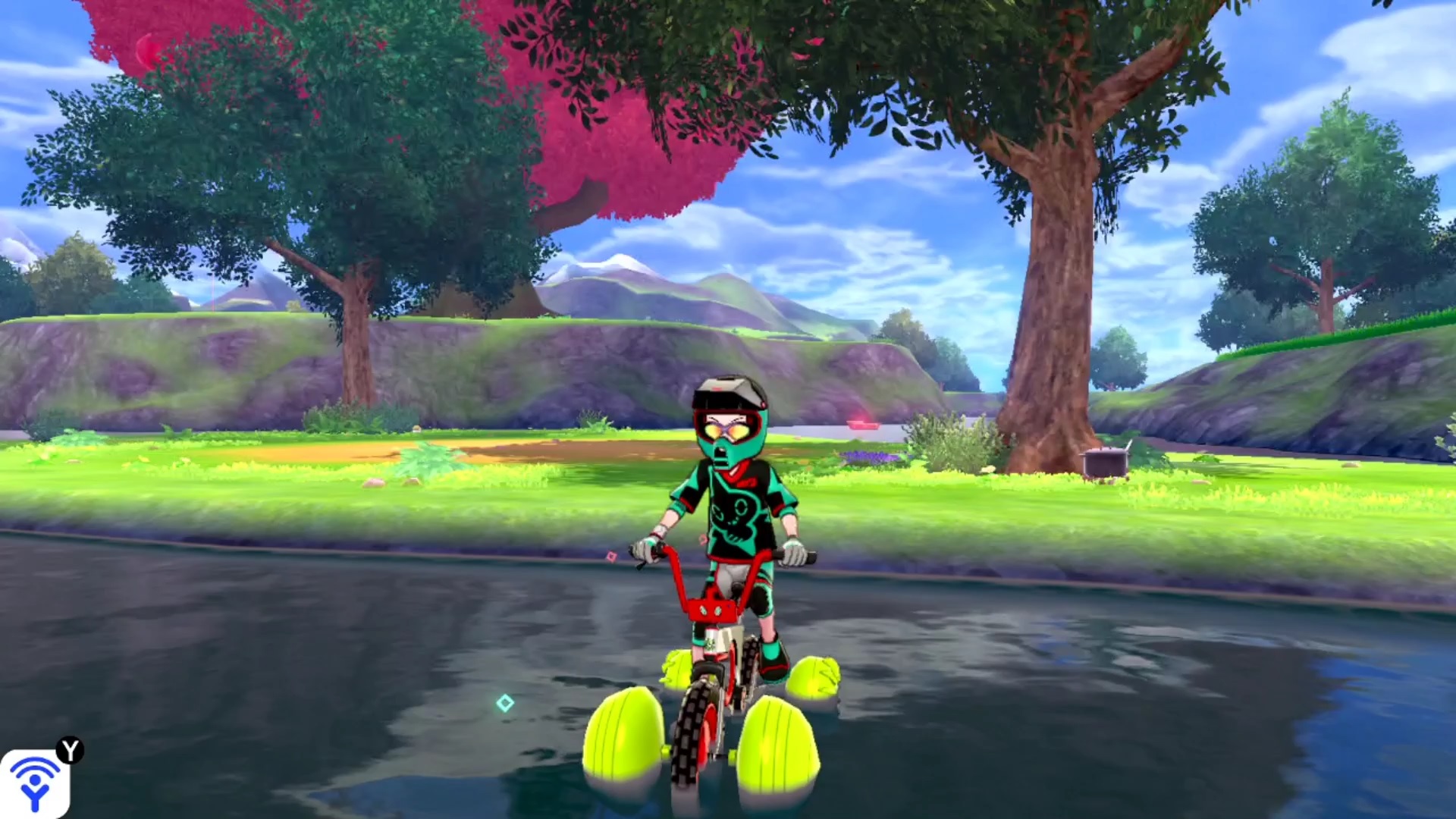 Pokémon Sword and Shield Keldeo: The Trainer riding a floating bike in water, with various trees and hills behind them.