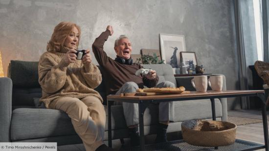 Two elderly people dressed in beige sit on a sofa holding a gamepad. The man cheers, the woman claps