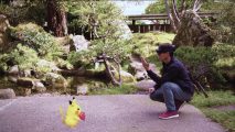 A man squats down wearing chunky VR glasses, Pikachu eats a berry in front of him. A real garden is the background