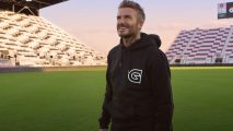 David Beckham, standing in the middle of a football field, wearing a black Guild hoodie