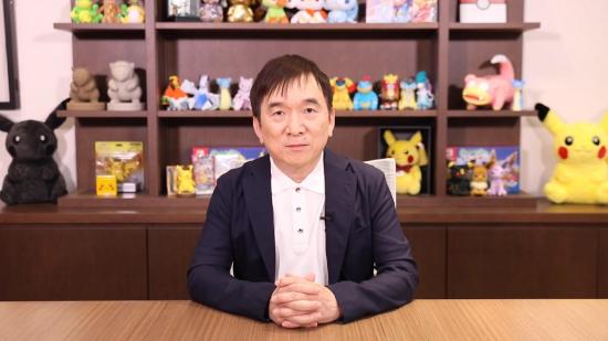 President Ishihara of the Pokémon Company sits at a desk staring at the camera, Pokémon plushies line the shelves behind him