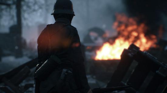 A soldier stands in front of the camera looking across the battlefield with a fire in the distance.