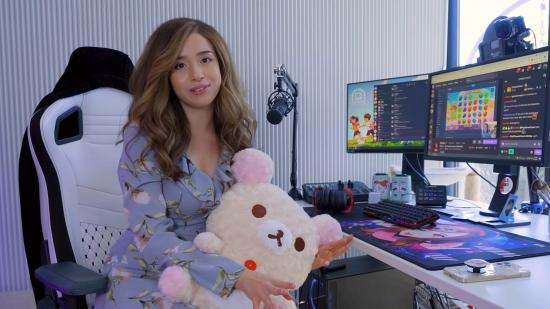 Livestreamer Pokimane sitting at her streaming set up. She is wearing a lilac dress and holding a large soft toy resembling a bear. On her desk is a streaming mic and two PC monitors