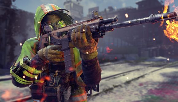 XDefiant Ubisoft Rainbow Six Siege: A person in a green hooded coat, with small flames coming of their body, firing a large assault rifle