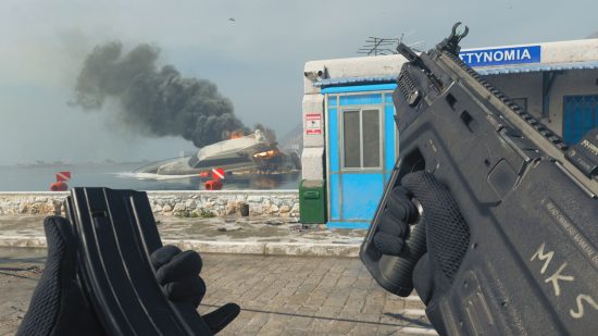MW3 meta: The player reloading the RAM-7 assault rifle in a coastal town, with a crashed plane in flames in the distance.