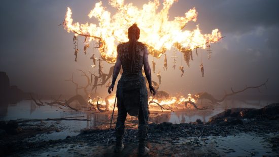 Best Xbox games: Senua standing with her back facing the camera. In the distance is a large tree with hanging bodies set on fire.
