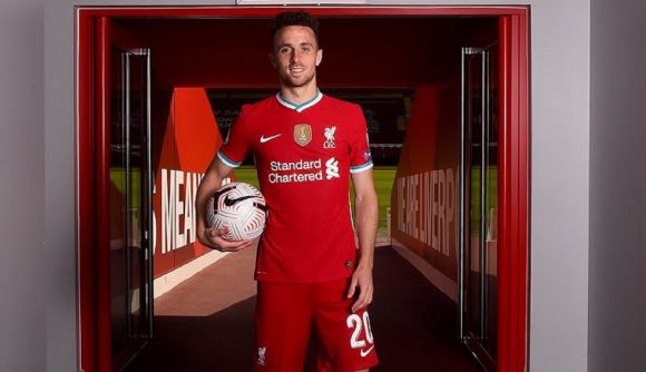 Diogo Jota stands in a red Liverpool kit, holding a ball under his arm and smiling at the camera