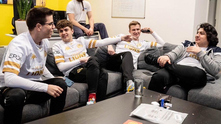 A group of young men in white jerseys with gold details. There is a table with a glass of water, lip balm, airpods, paper, and a wallet on