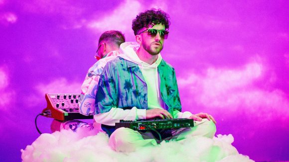 Two men play synthesisers sat on a fake cloud, with a purple sky behind them